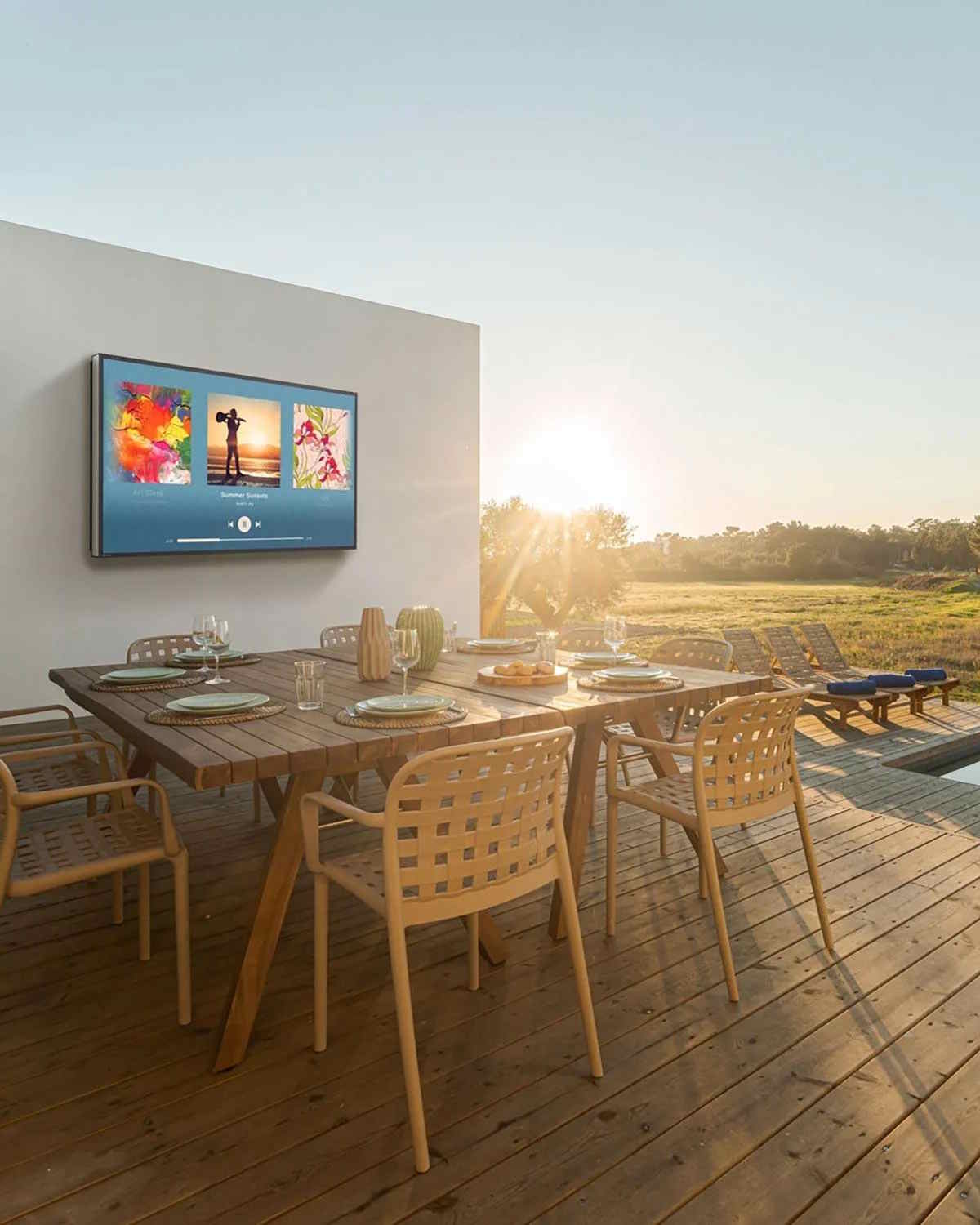 Outdoor dining setup on a wooden deck with a table set for six, a wall-mounted TV displaying music visuals, and sunset views over a grassy landscape.