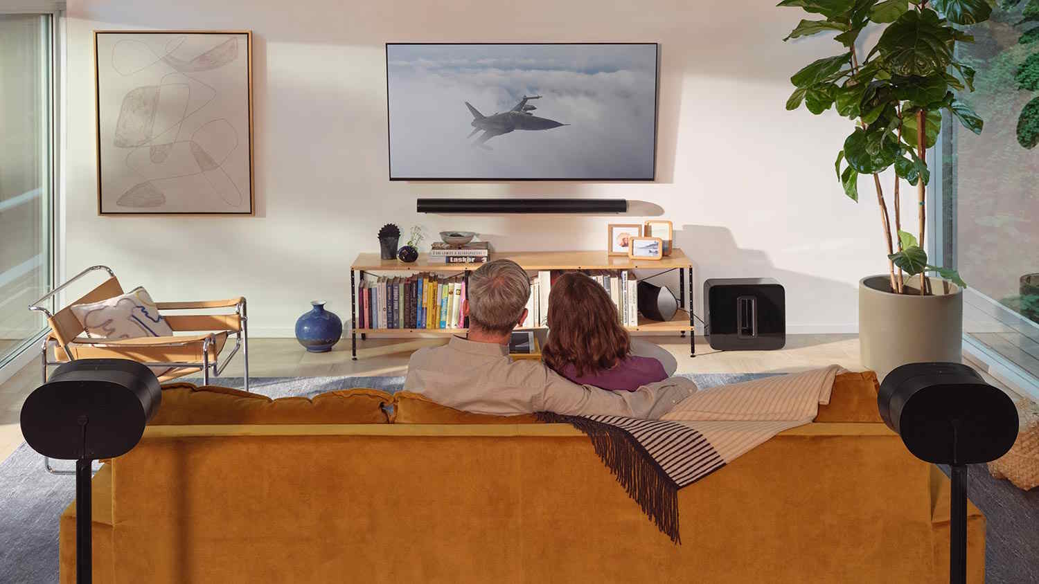 Couple sitting on a mustard yellow couch watching a jet scene on a wall-mounted TV, surrounded by bookshelves, plants, and surround sound speakers.