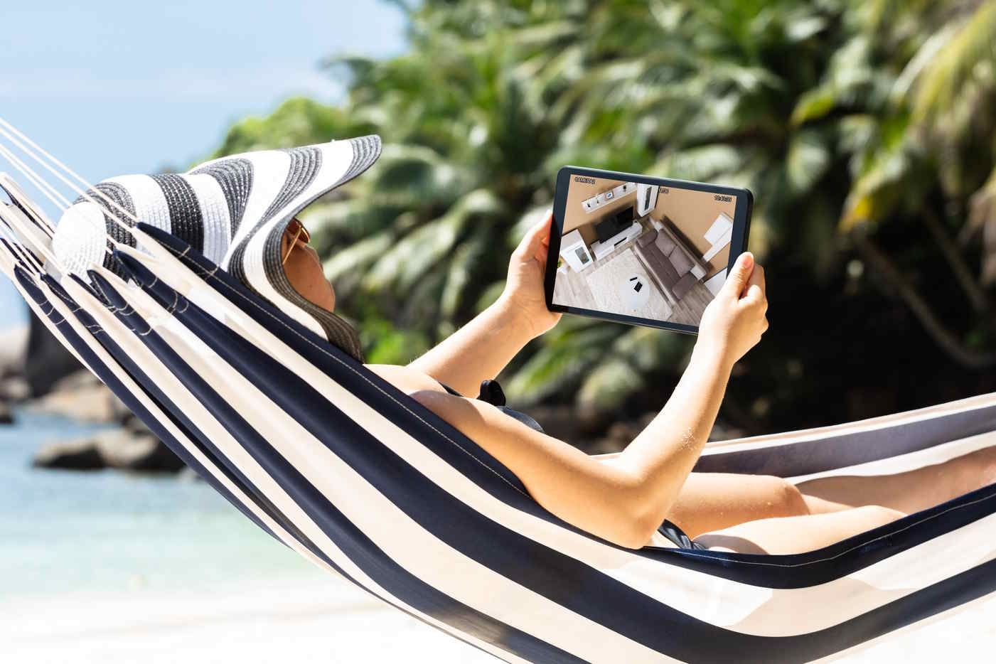 Woman relaxing on a striped hammock at the beach, viewing a home interior on a tablet.