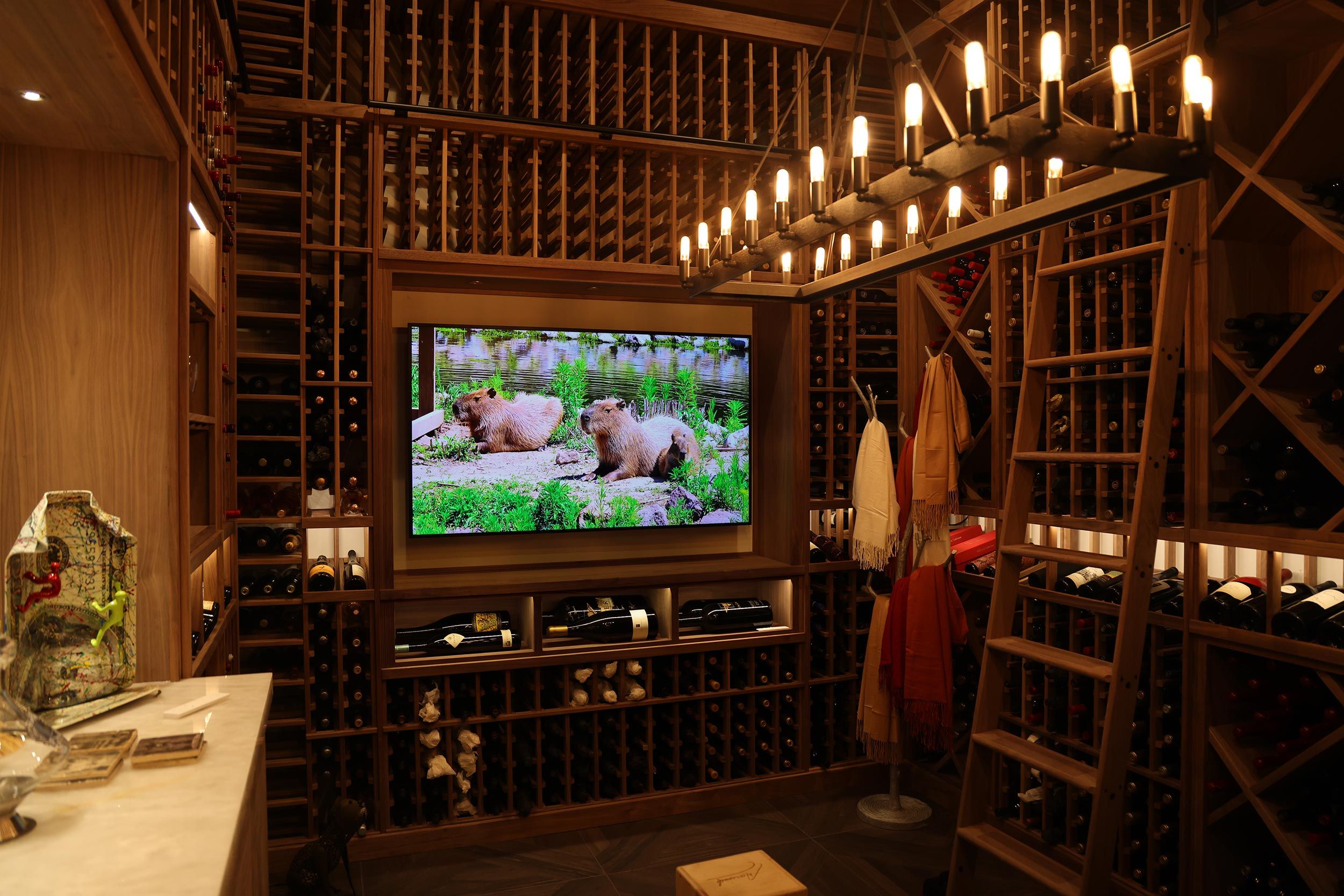 A luxurious wine cellar with custom wooden racks, a central TV display, and warm chandelier lighting.