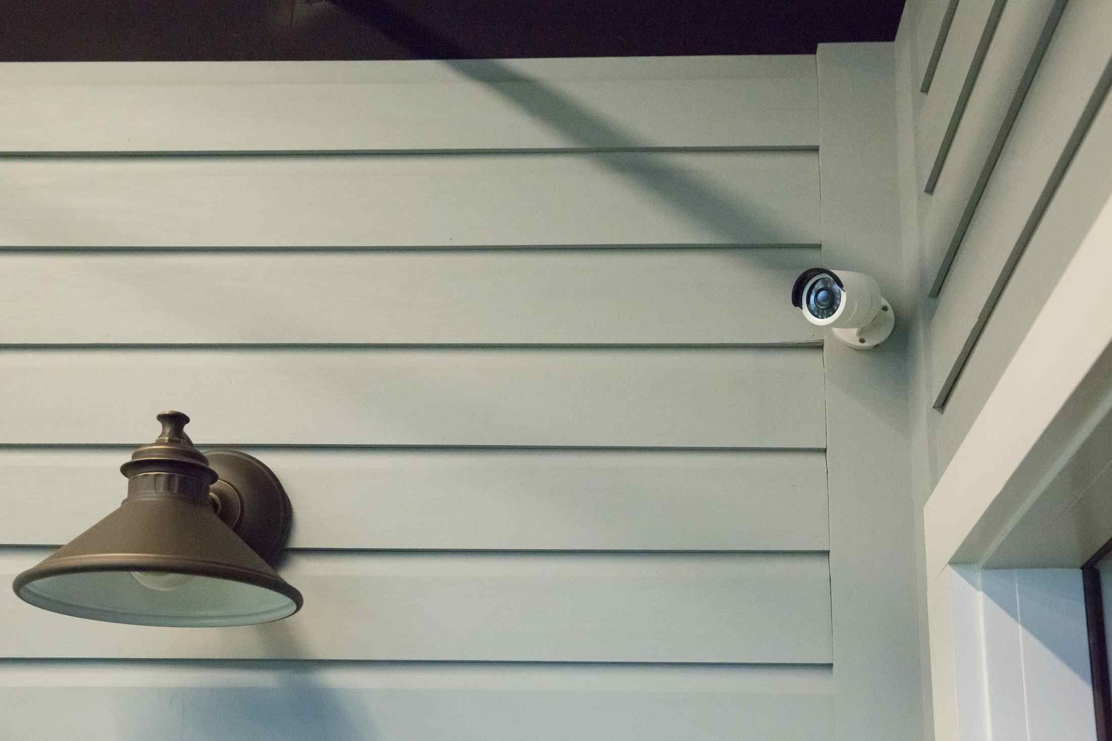 Outdoor security camera mounted on light gray siding near a wall-mounted bronze light fixture.
