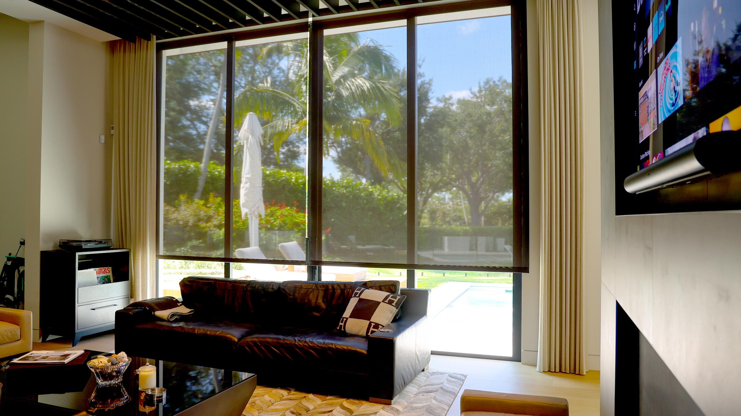 A modern living room with large glass doors featuring motorized shades overlooking a pool area.