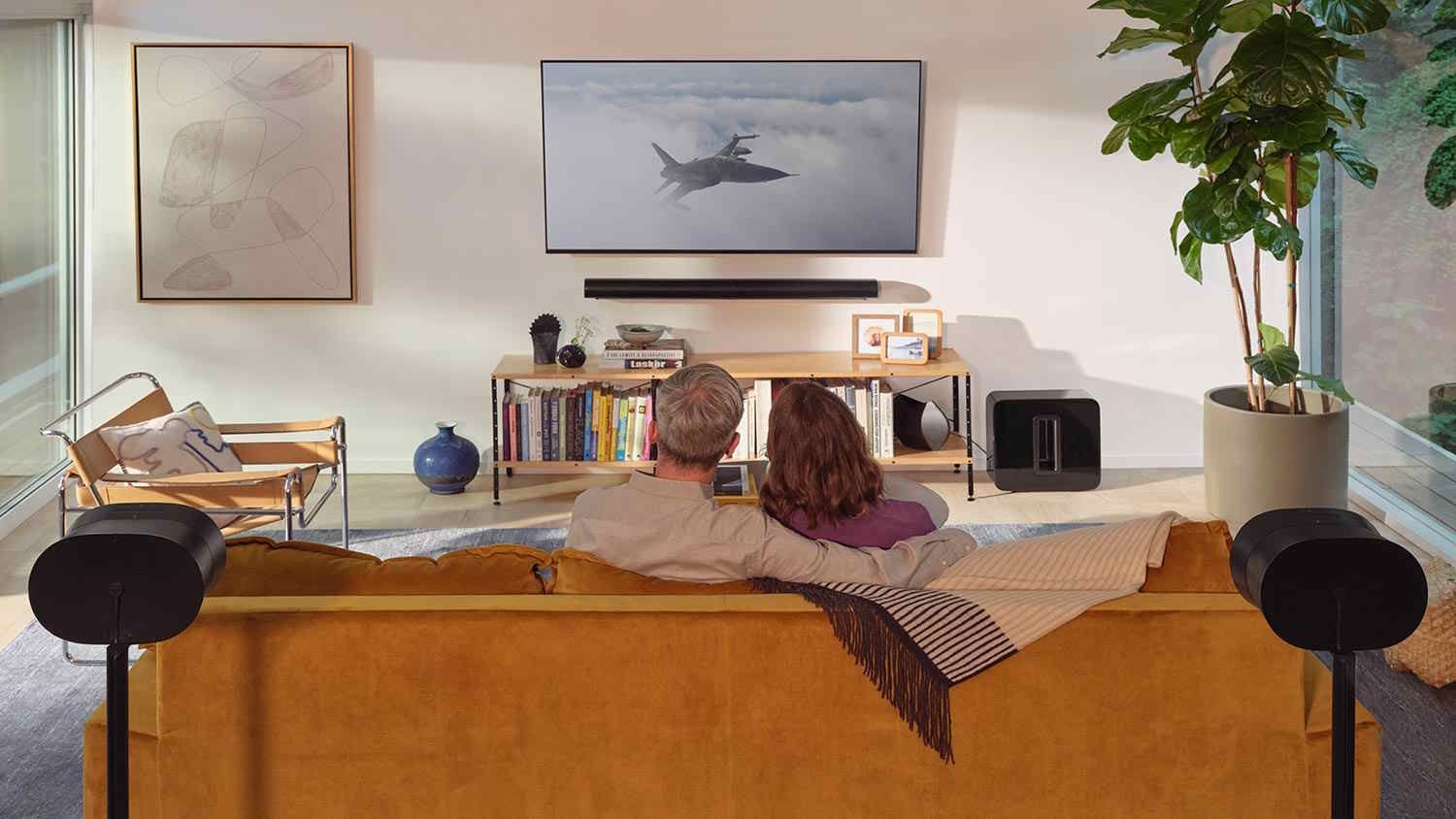 Couple sitting on a mustard yellow couch watching a jet scene on a wall-mounted TV, surrounded by bookshelves, plants, and surround sound speakers.