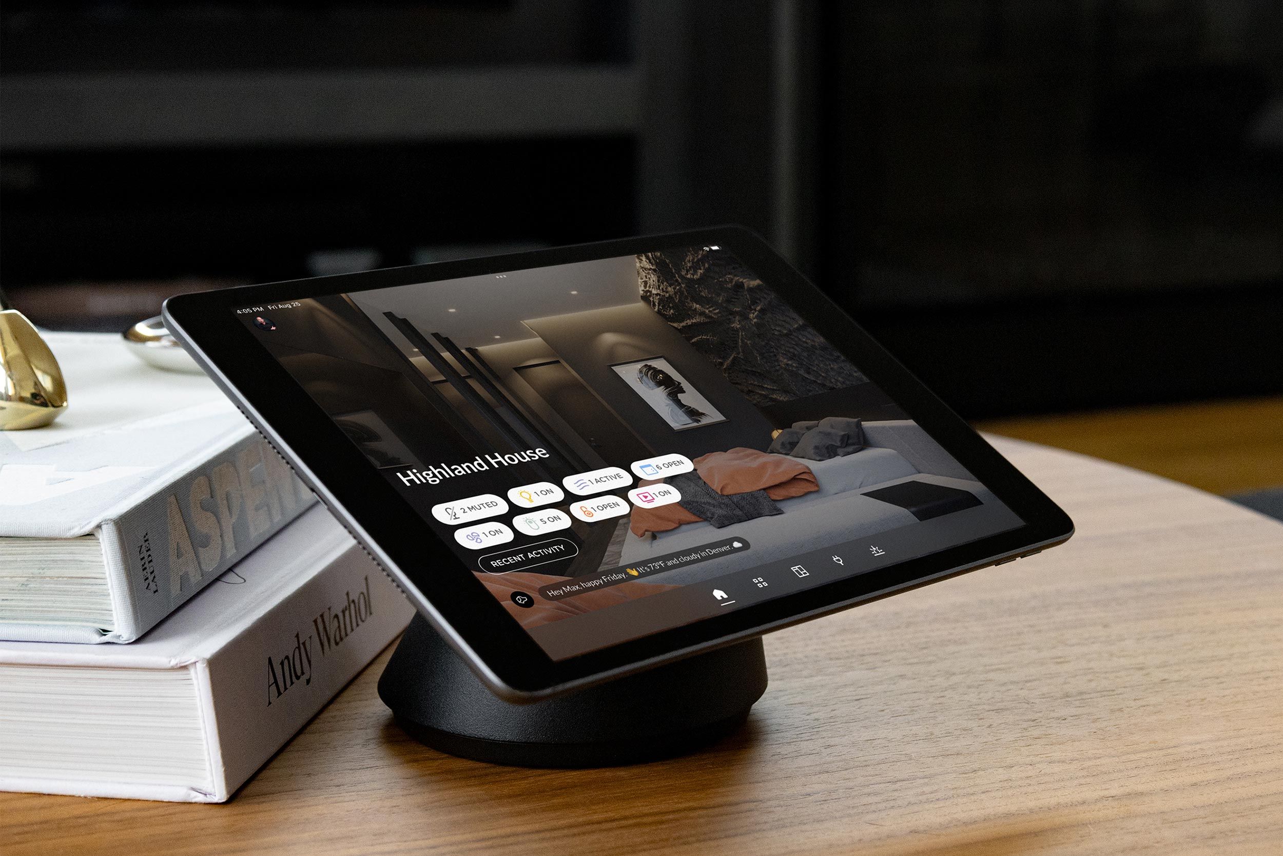 Tablet on a stand displaying a smart home control dashboard for “Highland House,” placed on a wooden table next to stacked books.